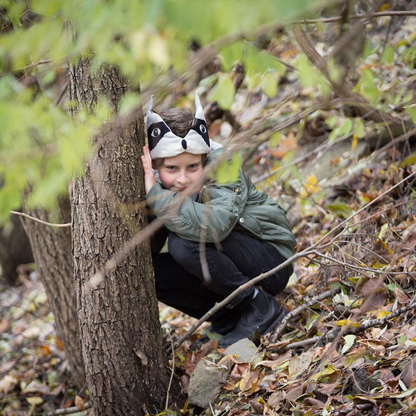 A child wearing a fabric racoon headdress playing in the woods, crouched behind a tree.